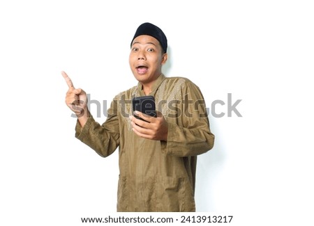 surprised asian muslim man wearing islamic clothes pointing to above with holding mobile phone isolated on whtie background