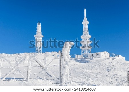 Frozen television or cellular tower in heavy snow near ski center. Telecommunication towers with dish and mobile antenna against blue sky in winter mountains.