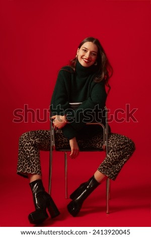  Fashionable happy smiling woman wearing trendy green turtleneck sweater, leopard print jeans, black leather platform chunky heel boots, posing on red background. Full-length studio fashion portrait