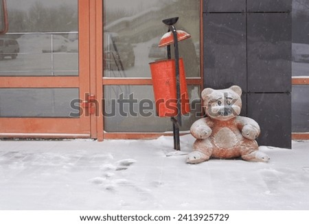 Teddy bear sitting in the snow near the building. Abandoned toy bear alone on the street in winter. A snow-covered freezing teddy bear near the urn. Royalty-Free Stock Photo #2413925729