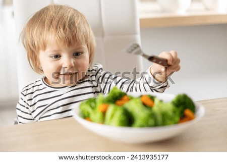Cute little girl is eating broccoli and carrot vegetables with a fork.