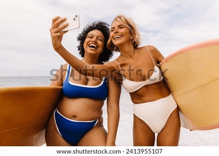 Happy surfing friends capturing their beach vacation moments with a selfie, embodying the shared joy of riding the waves. Two young women in bikinis enjoying beach activities in summer.