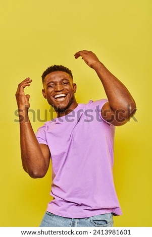 excited and trendy african american man in violet t-shirt gesturing laughing on yellow backdrop