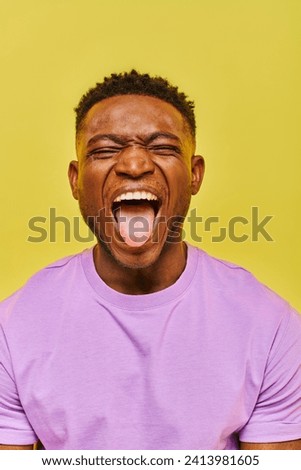 young african american man with closed eyes grimacing and sticking out tongue on yellow backdrop