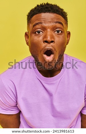 amazed african american man in purple t-shirt with open mouth looking at camera on yellow backdrop
