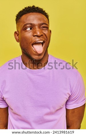 funny african american man in purple t-shirt sticking out tongue and looking away on yellow backdrop