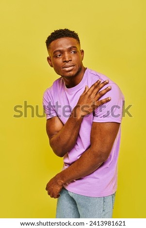 young handsome african american man in violet t-shirt posing and looking away on yellow backdrop