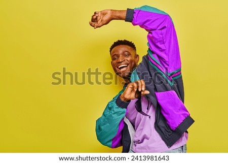 carefree african american man in stylish jacket holding sunglasses and dancing on yellow backdrop