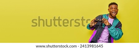 smiling african american man in bright jacket holding sunglasses and dancing on yellow, banner