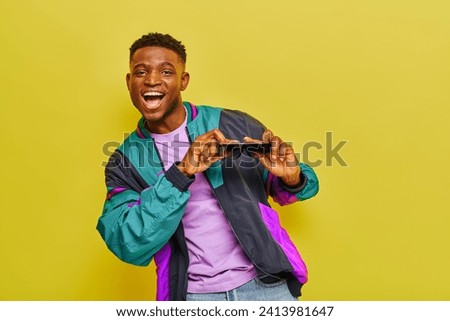 overjoyed african american man in bright jacket holding sunglasses and dancing on yellow backdrop
