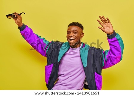 excited african american man in bright jacket holding sunglasses and dancing on yellow backdrop