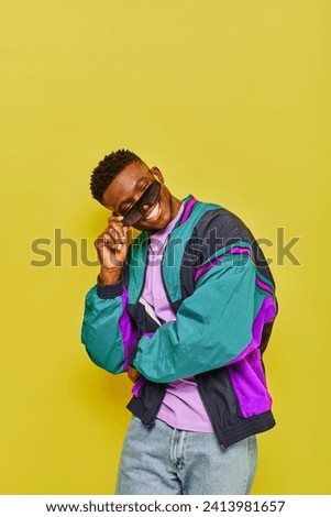 joyful african american man in colorful windbreaker jacket looking over trendy sunglasses on yellow