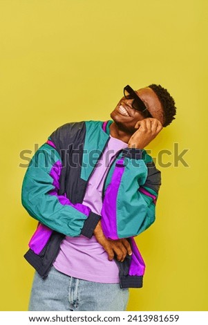 cheerful stylish african american guy in bright windbreaker jacket and sunglasses smiling on yellow