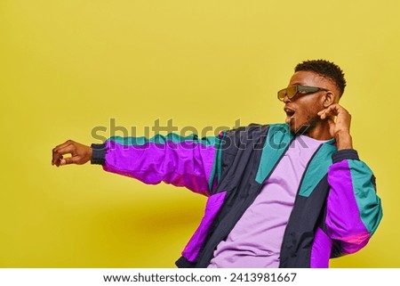 fashionable and energetic african american man in bright jacket and sunglasses dancing on yellow