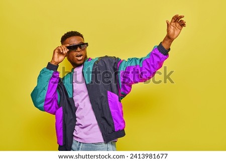 excited african american man in sunglasses and trendy colorful jacket waving hand on yellow