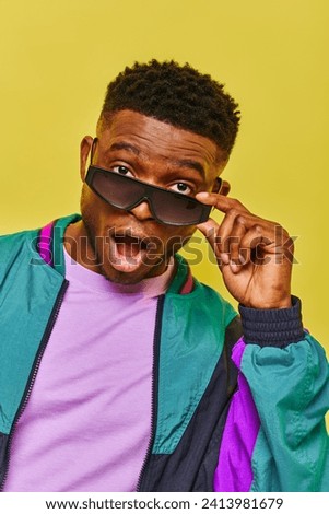 impressed african american man with open mouth holding sunglasses and looking at camera on yellow