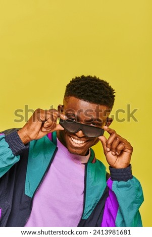 cool african american man in fashionable sunglasses and colorful jacket smiling at camera on yellow