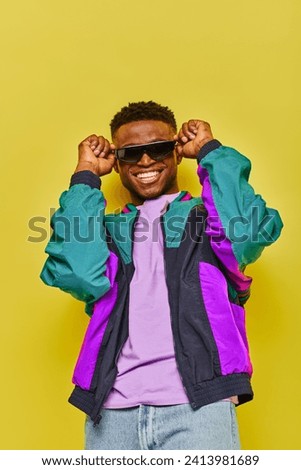 overjoyed african american man in bright jacket and trendy sunglasses smiling at camera on yellow