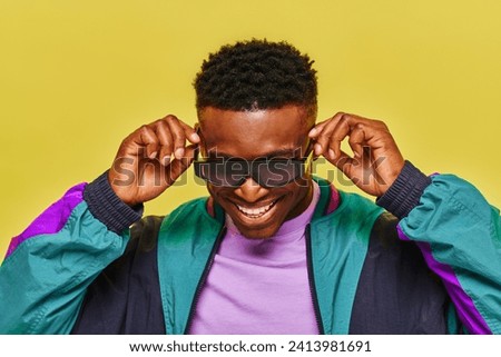 stylish african man in colorful jacket adjusting sunglasses and smiling on yellow backdrop