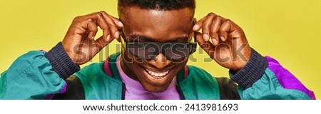 trendy african guy in bright jacket adjusting sunglasses and smiling on yellow backdrop, banner