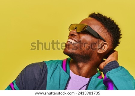 carefree african american man in windbreaker jacket and stylish sunglasses looking away on yellow