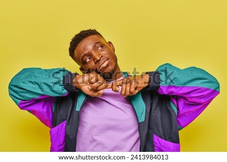 dreamy positive african american man in colorful windbreaker jacket looking away on yellow backdrop
