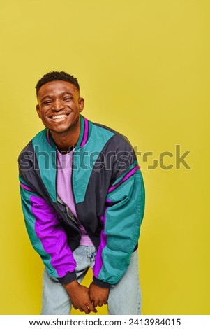 overjoyed trendy african american man in bright windbreaker smiling and posing on yellow backdrop