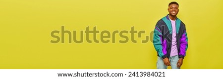 joyful african american man in colorful windbreaker smiling and posing on yellow background, banner