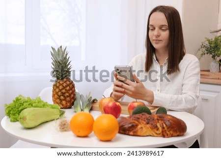 Beautiful Caucasian woman wearing white shirt cooking vegetables in cozy light kitchen while looking recipe on smartphone browsing website on tutor cooking class and smiling
