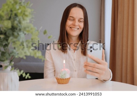 Smiling cheerful Caucasian woman with birthday cake and smartphone in home interior celebrating her holiday with her friend online via video call looking at mobile phone screen
