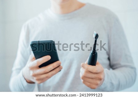 Close up man puts settings of his electronic toothbrush with mobile phone app. Wireless connecting sonic toothbrush with smart phone app. Modern home health care technology concept. Selective focus Royalty-Free Stock Photo #2413999123