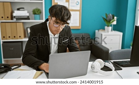 Serious, young hispanic teenager embracing technology, working as a business professional in the office. sitting at his desk, vested in success, he dives into work on his laptop
