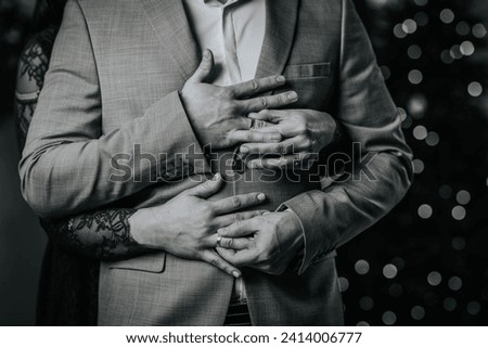 monochrome close-up of a couple's hands clasped around a man in a suit, with the woman's hand wearing a wedding ring. Royalty-Free Stock Photo #2414006777