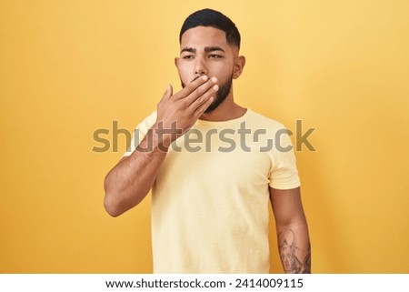 Young hispanic man standing over yellow background bored yawning tired covering mouth with hand. restless and sleepiness. 