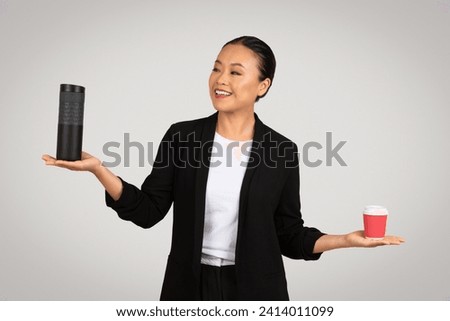 Smiling Asian businesswoman comparing a large black thermal tumbler in one hand with a small red disposable coffee cup in the other, suggesting choice or decision on a grey background