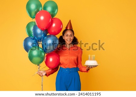 Cheerful African American teen girl posing with bunch of colorful balloons and birthday cake with candles, celebrating and having bday party over yellow studio background, smiling to camera
