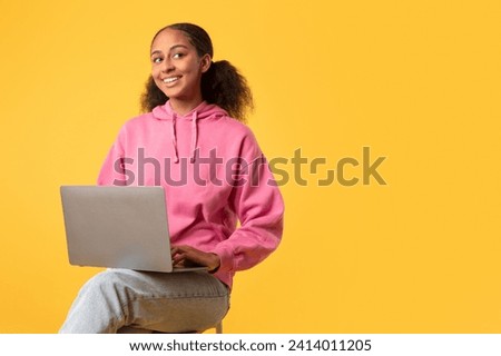 Black teen girl with notebook computer learning online sitting against yellow background, studio shot of digital native student studying with internet technology. E-learning offers, copy space