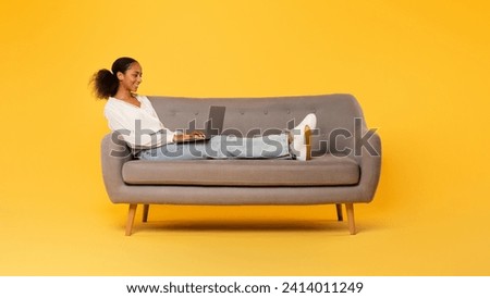 Smiling African American teen girl typing on laptop, using computer for e-learning, browsing educational websites while sitting on comfortable sofa against yellow studio backdrop. Panorama