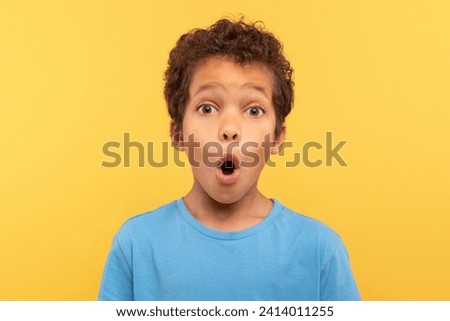 Astonished young boy with curly hair, mouth agape in surprise, wearing light blue shirt against plain yellow background, capturing moment of amazement Royalty-Free Stock Photo #2414011255