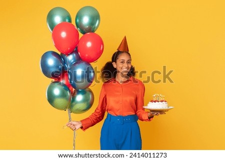 Cheerful black teenager girl posing with bunch of festive balloons and birthday cake, embodying celebration against yellow studio backdrop, smiling to camera. Bday holiday and party fun