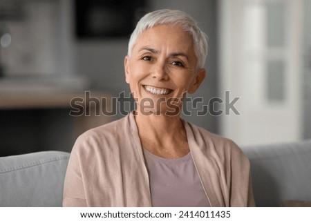 Portrait of happy healthy older woman with short haircut and gray hair posing for photo at home, smiling to camera with cheerful expression, sitting on comfortable couch at home. Royalty-Free Stock Photo #2414011435