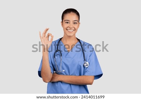 Positive caucasian millennial lady doctor or nurse in blue uniform show ok sign with hand, isolated on gray studio background. Professional health care approving, sale recommendation