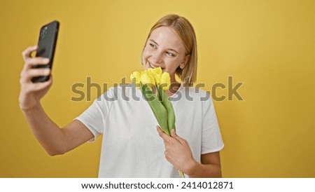 Young blonde woman holding bouquet of flowers making selfie by smartphone over isolated yellow background