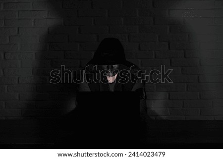 Hacker in black mask and hood at the table in front of the monitor
