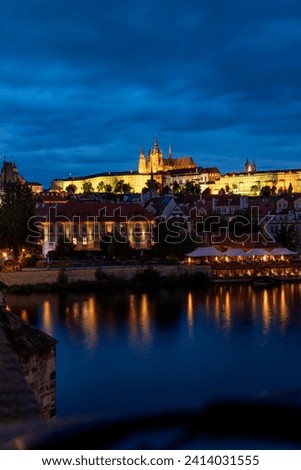 Nighttime photograph taken in Prague, Czech Republic, featuring a view of Prague Castle, the river, and a glimpse of an ancient bridge, illuminated by the city lights