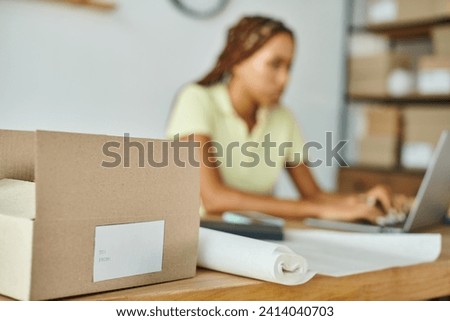 focus on cardboard box with label with blurred african american female merchant on background
