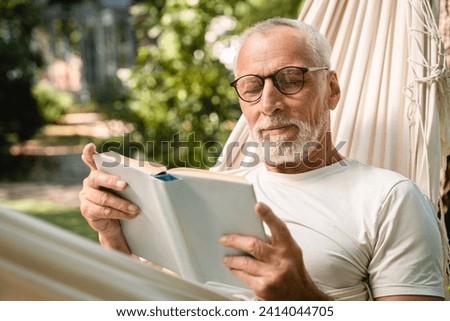 Concentrated dreamy old senior elderly caucasian grandfather man relaxing resting in hammock while reading book outdoors in park garden forest Royalty-Free Stock Photo #2414044705