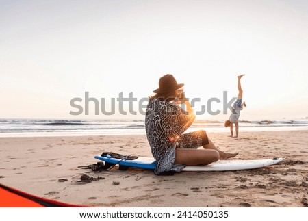 Young woman sitting on surfboard- taking pictures of young man- practicing handstands on the beach