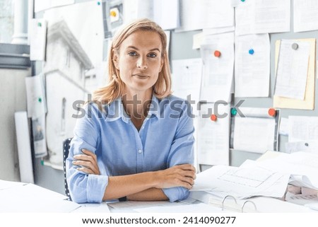 Portrait of confident woman sitting at desk in office surrounded by paperwork Royalty-Free Stock Photo #2414090277