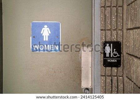 Two women's restroom signs on a concrete wall, one standard blue and one black with accessibility symbol.